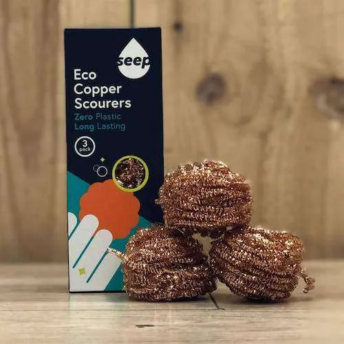 Three copper scouring balls next to their dark blue Seep Eco Copper Scourers box on a wooden surface with a wooden background.