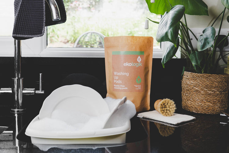 Brown pouch with a green stripe along the top. Wording washing up pods. White bowl filled with white dishes and white bubbles in sink.