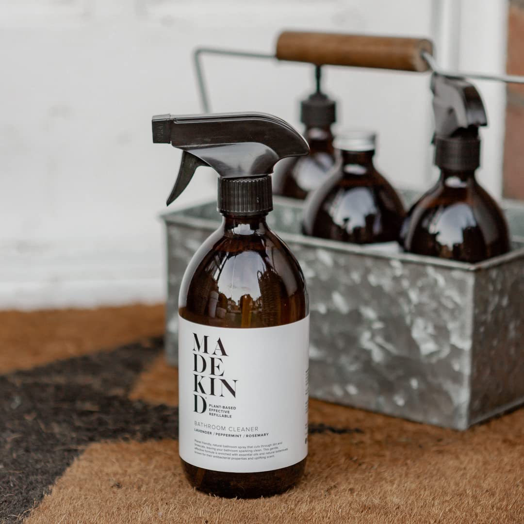 A brown glass spray bottle labeled 'MADE IN KIN D RAINBOW CLEANER' stands in front of a galvanized metal caddy holding two more spray bottles, set on a textured brown surface against a light neutral background.