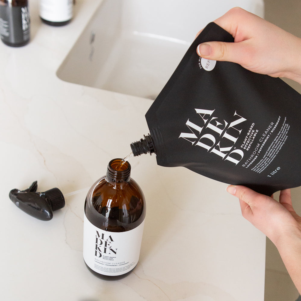 Person pouring liquid from a black 'MADE IN' bag into an amber bottle on a bathroom counter.
