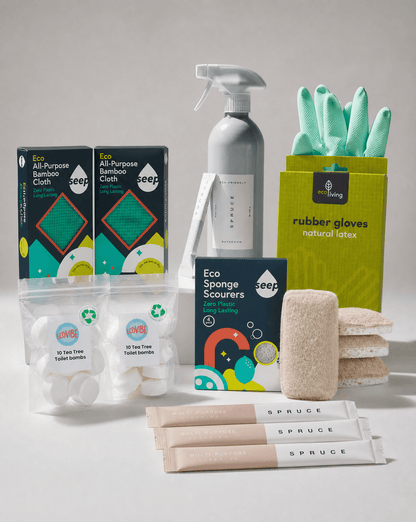 A coordinated set of eco-friendly cleaning and bath products arranged on a light gray studio background. Visible items include two black-and-teal Seep bamboo cloth boxes, two clear bags of toilet paper, a gray spray bottle, a green box of rubber gloves, a box of sponge scours, a beige scrubber, and three pink-and-white SPRUCE hand towels.