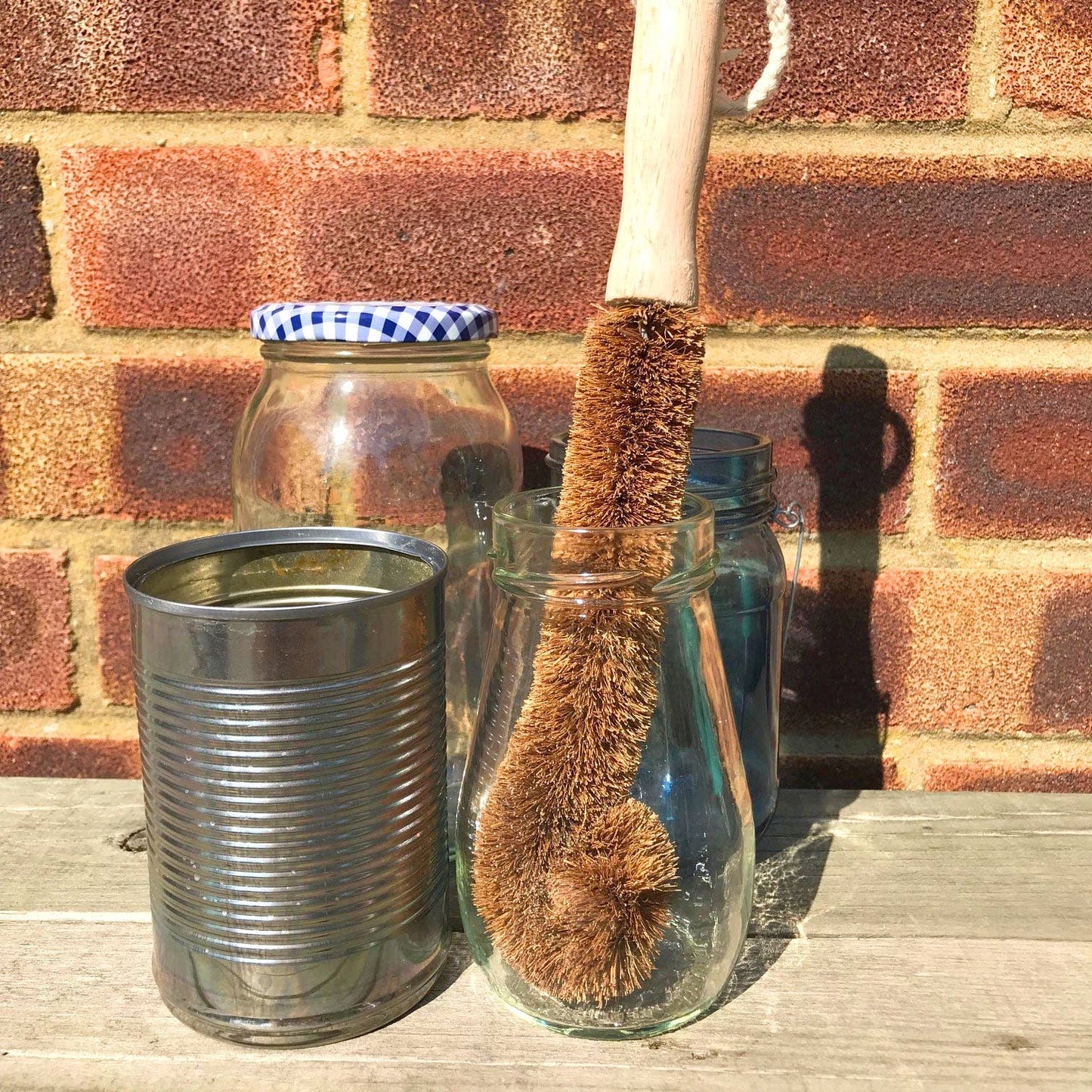 A natural brown coconut scrubber with a light wood handle stands in a clear glass jar, next to a metal tin can and a glass jar with a blue-and-white check lid, set against a brick wall on a wooden surface.