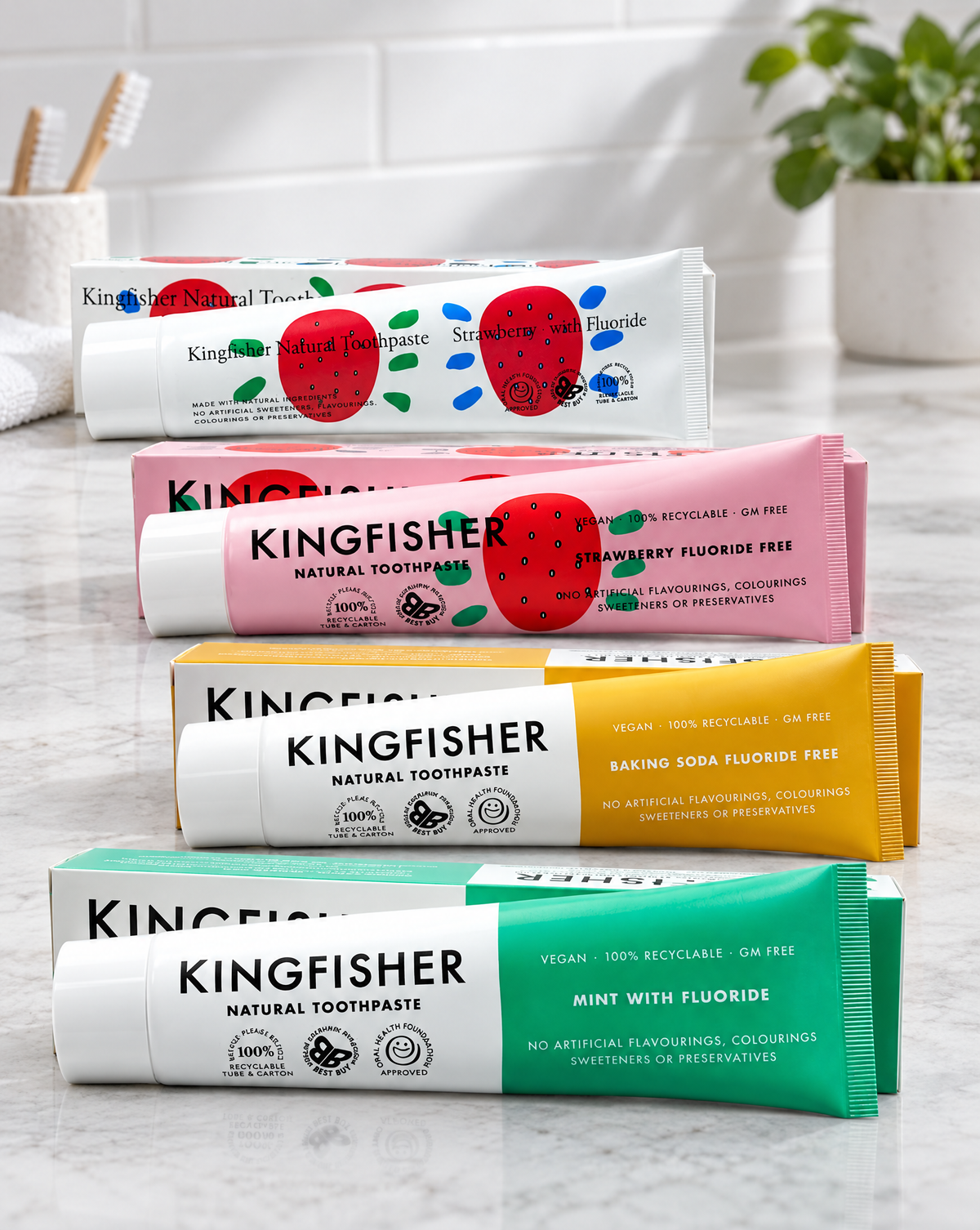 Five tubes of Kingfisher Natural Toothpaste arranged diagonally on a light marble surface. The toothpaste tubes are white, pink, yellow, and green, decorated with illustrations of red strawberries and flavor labels including Strawberry Fluoride Free, Baking Soda Fluoride Free, and Mint with Fluoride. Packaging recyclable.  Free from additives.