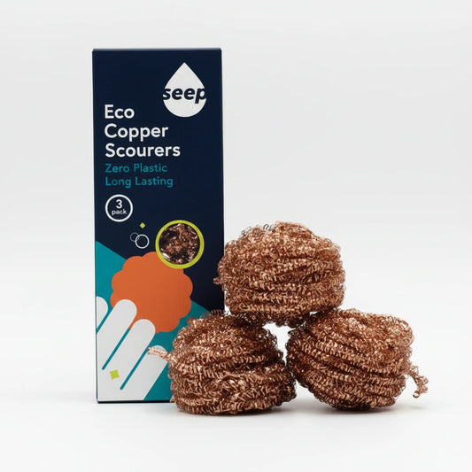 Three copper scouring balls next to their dark blue Seep Eco Copper Scourers box on a wooden surface with a wooden background.