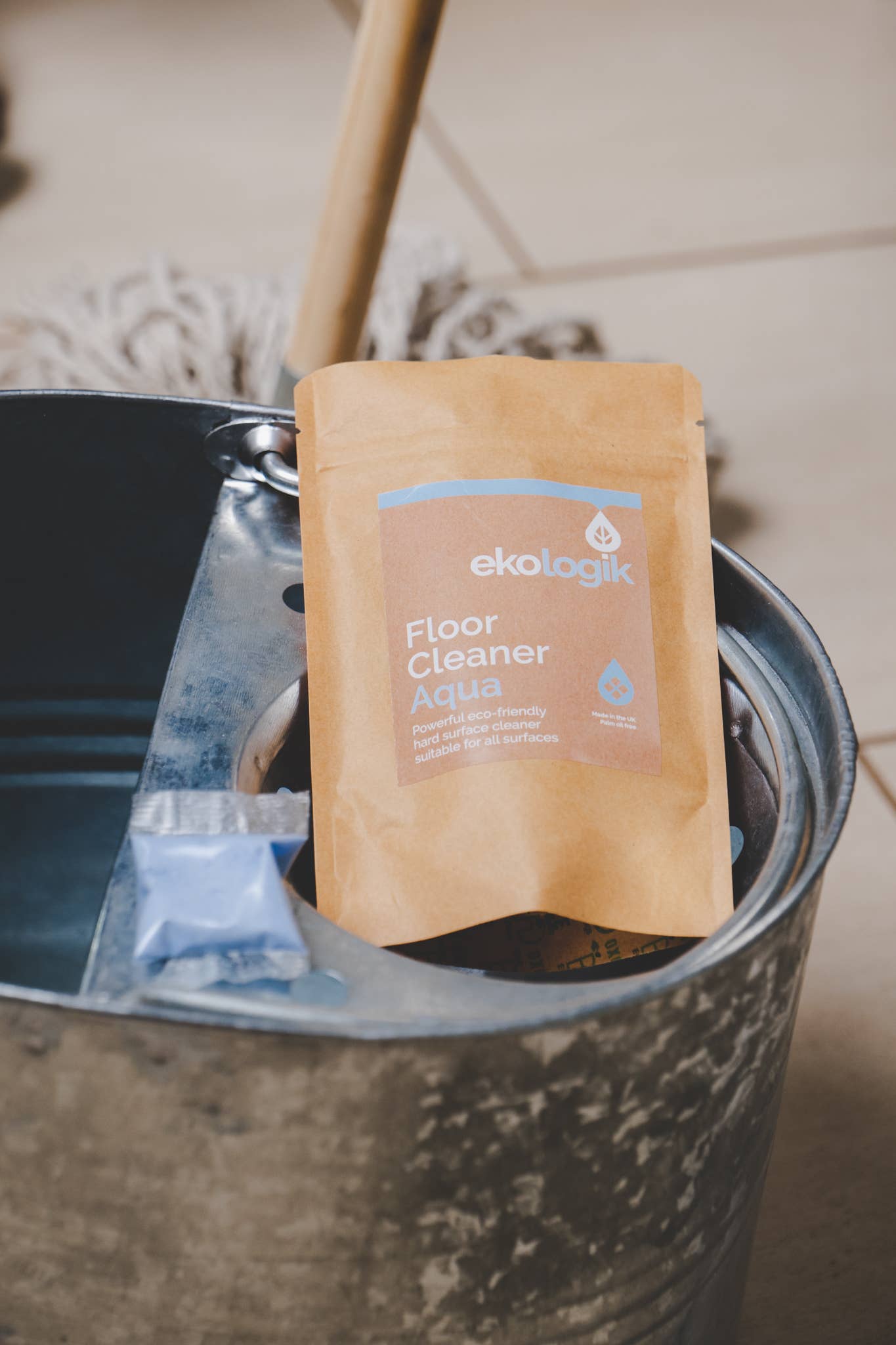pouch of ecologik aqua floor cleaner pods with one light blue pouch. Both on a metal mop bucket with mop in the background mopping cream floor tiles