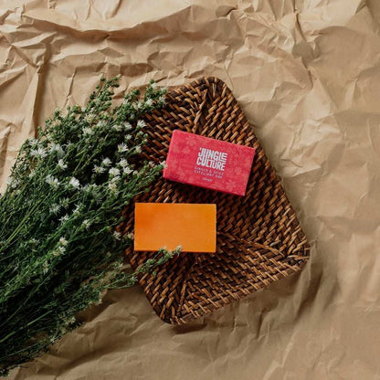 A styled flat-lay of a bar of soap in an orange container next to a pink box labeled 'JUNGUE CULTURE', placed on a brown woven mat over a crumpled brown paper background with dried green and white flowers.