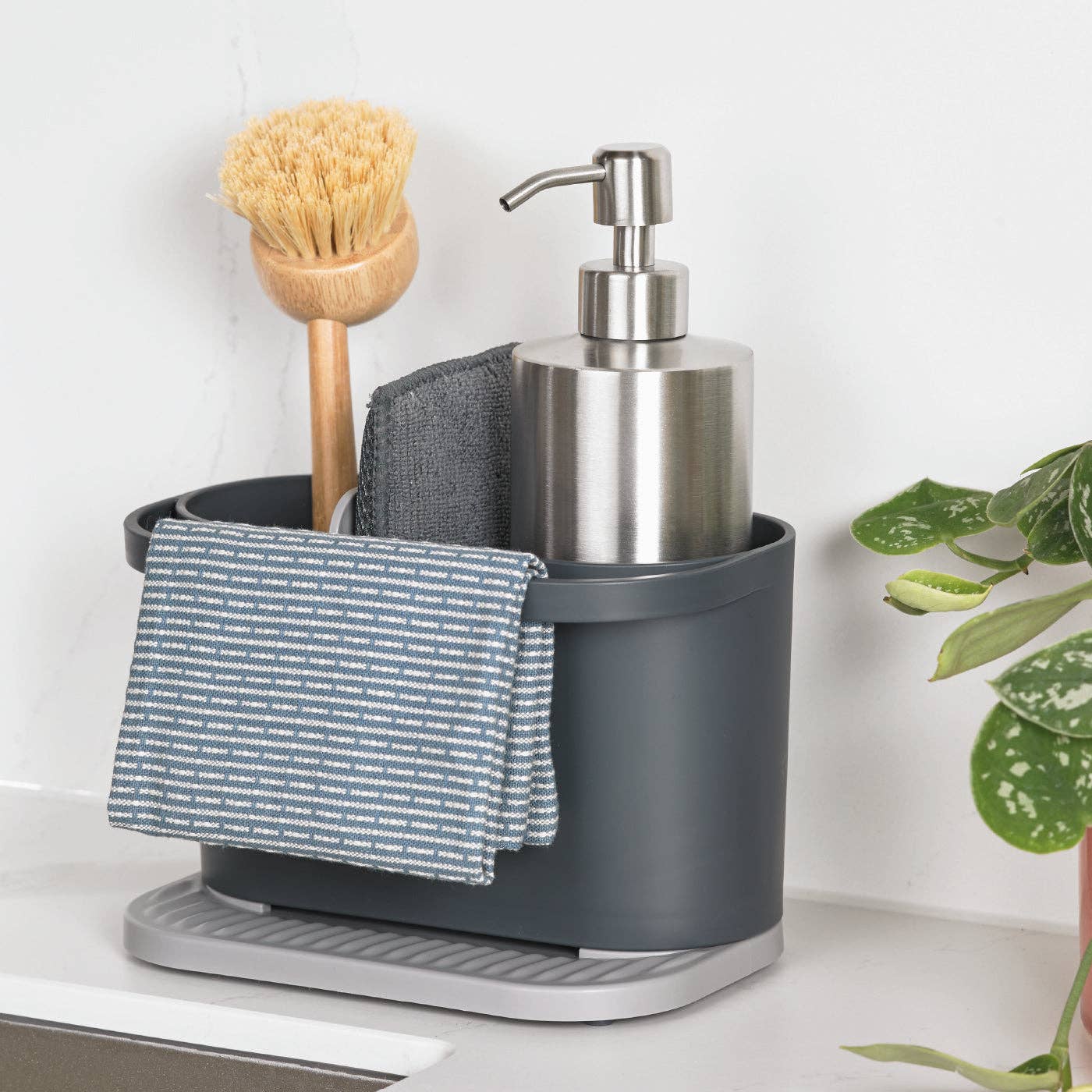Gray recycled plastic sink caddy with soap dispenser, scrub brush, and cloth on a white surface with a plant in the background