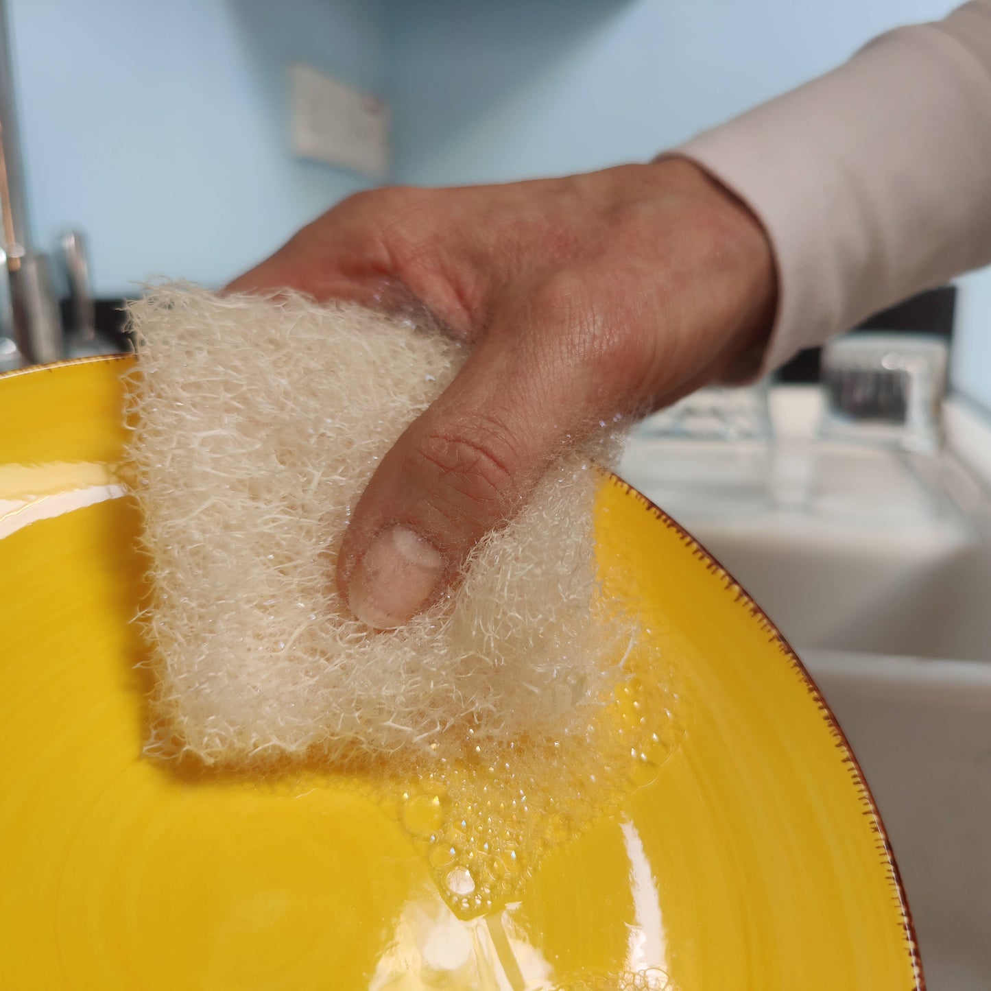 Hand holding a plastic free loofah dish sponge over a yellow plate with soap bubbles.