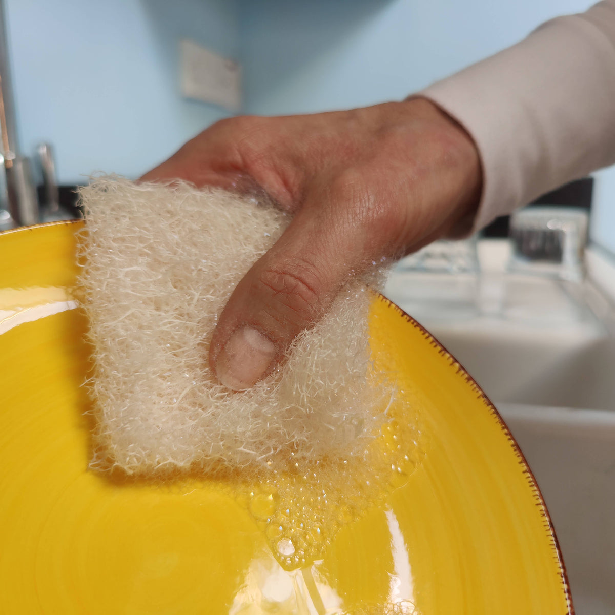 Hand holding a plastic free loofah dish sponge over a yellow plate with soap bubbles.