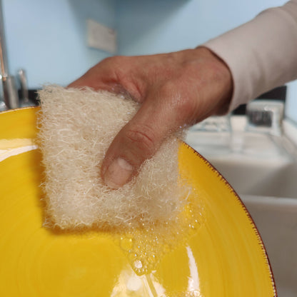 Hand holding a plastic free loofah dish sponge over a yellow plate with soap bubbles.