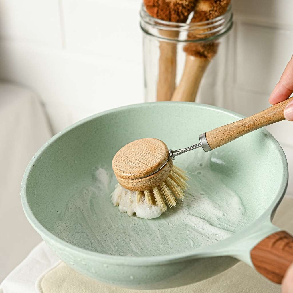 Person using a wooden scrubber to clean a green bowl with soap suds.