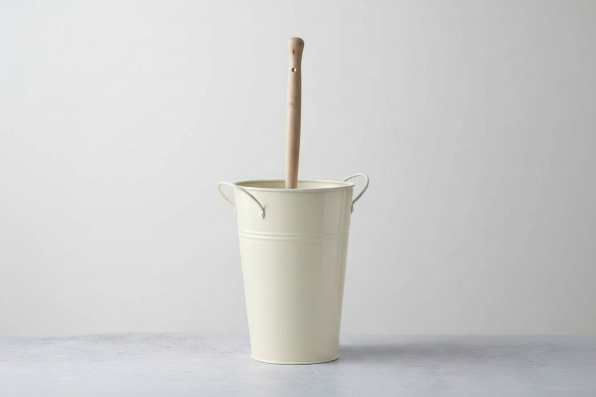 Beige metal bucket with a wooden handle against a white brick wall.  Plastic-free alternative