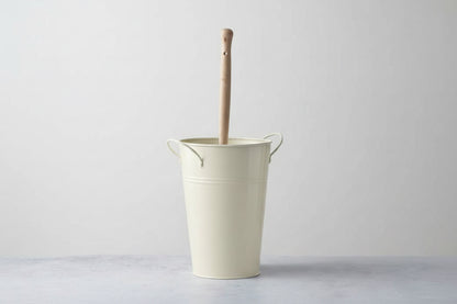 Beige metal bucket with a wooden handle against a white brick wall.  Plastic-free alternative