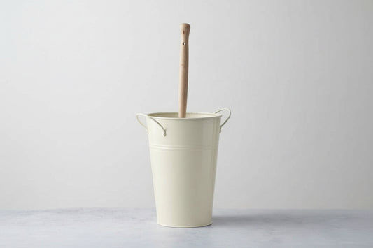 Beige metal bucket with a wooden handle against a white brick wall.  Plastic-free alternative