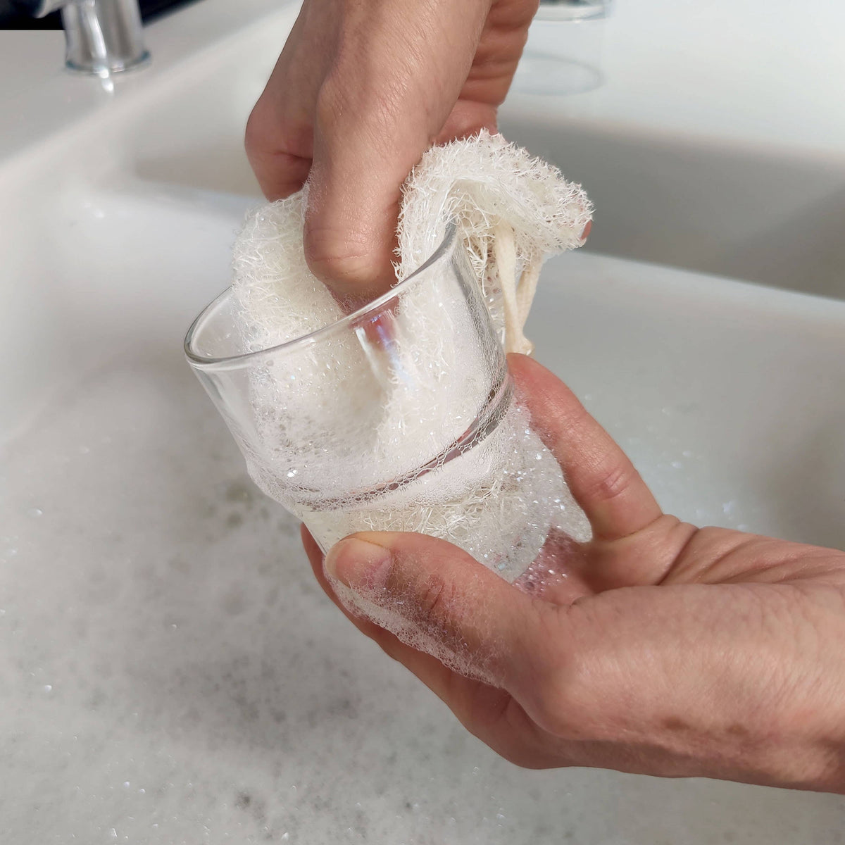 A close-up of hands washing a clear glass cup with a white, natural loofah plastic-free dish sponge