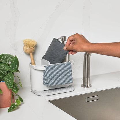 A white upcycled sink organiser on a kitchen sink, holding a grey sponge and a blue-and-white striped cloth, with a hand placing the sponge into the rack. A plant and faucet are visible in a real kitchen setting.